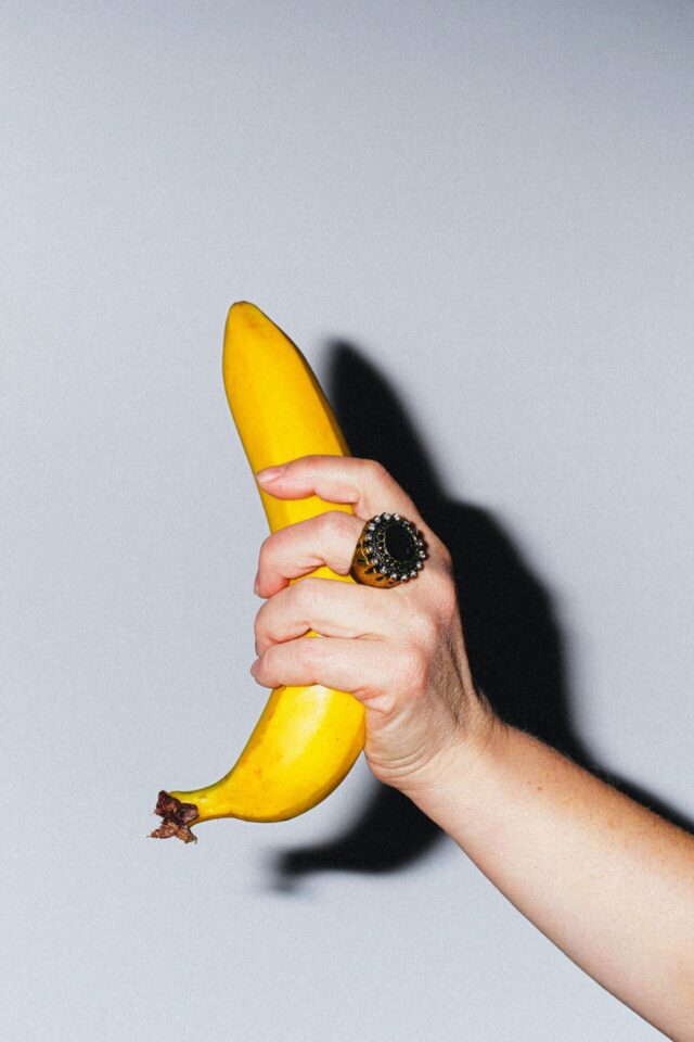 A vibrant yellow banana held in a hand with a decorative ring, staged against a neutral background.