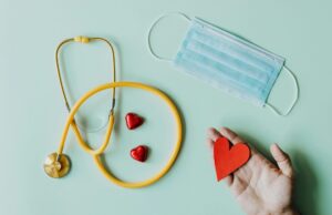 «La enfermería no es solo ciencia, es un arte de amor y paciencia» Top view of crop anonymous person hand with red paper heart on table with stethoscope and medical mask for coronavirus prevention